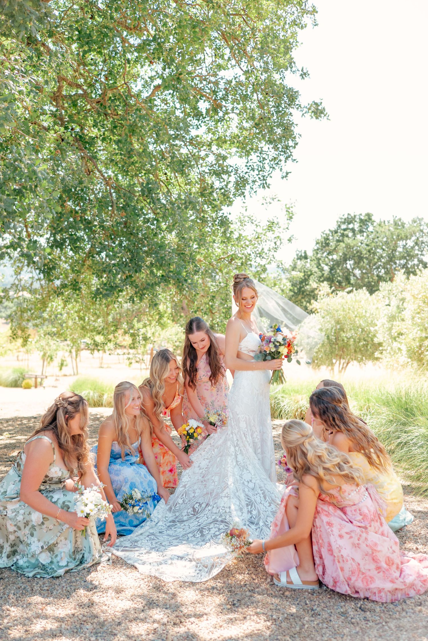 Bridesmaids in colorful dresses gather around a bride's dress on a sunny outdoor day.