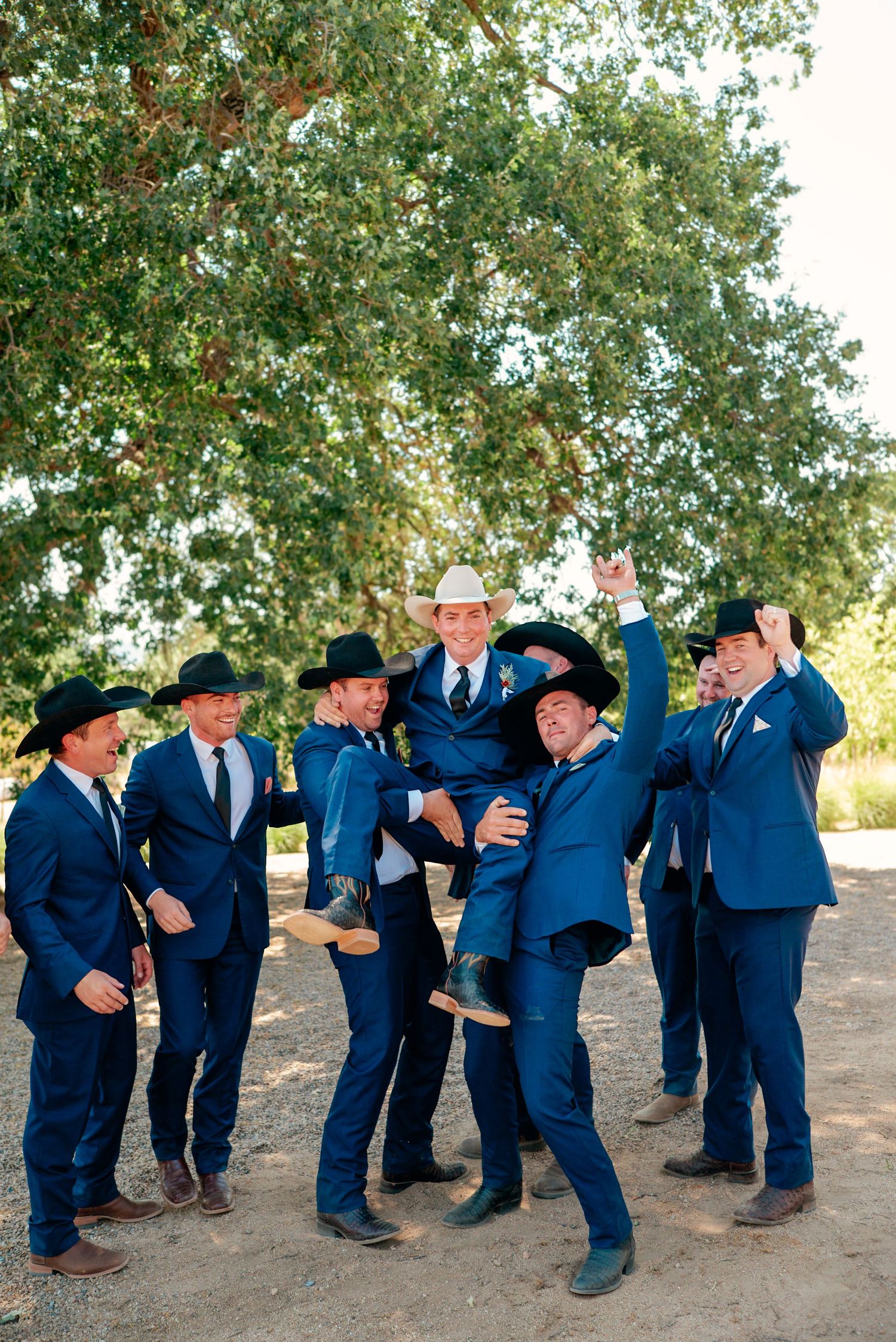 Groomsmen in matching navy suits and cowboy hats celebrate playfully outdoors under a tree.