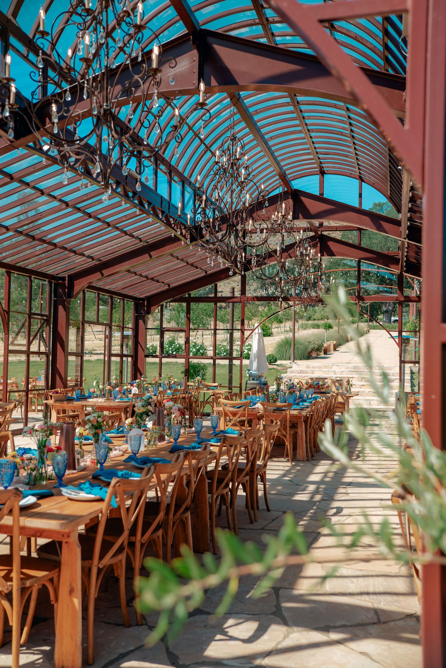 Elegant greenhouse reception venue with wooden tables and chairs set beneath a glass ceiling.
