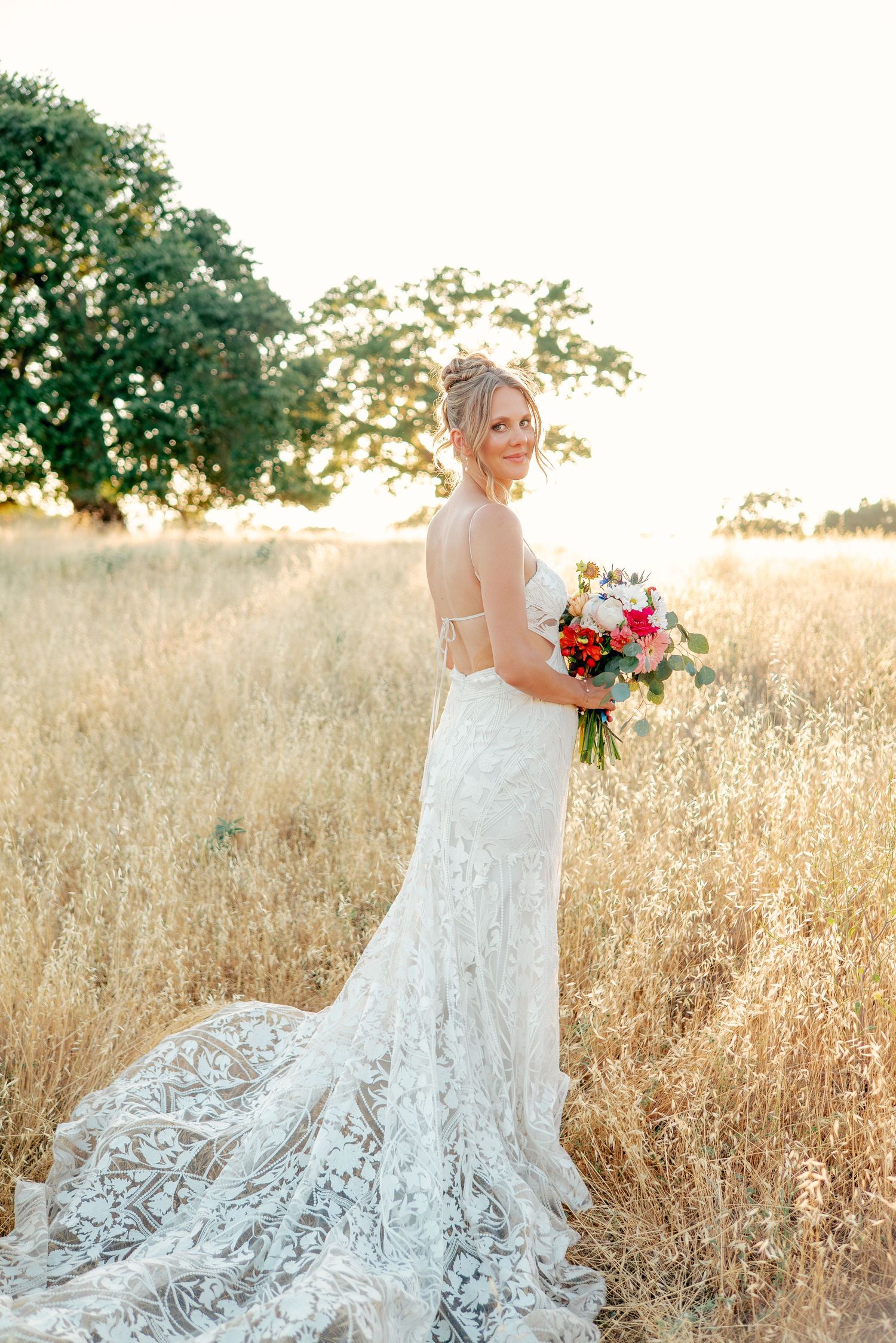 A bride in a lace wedding dress poses in a golden field at sunset holding a colorful bouquet.