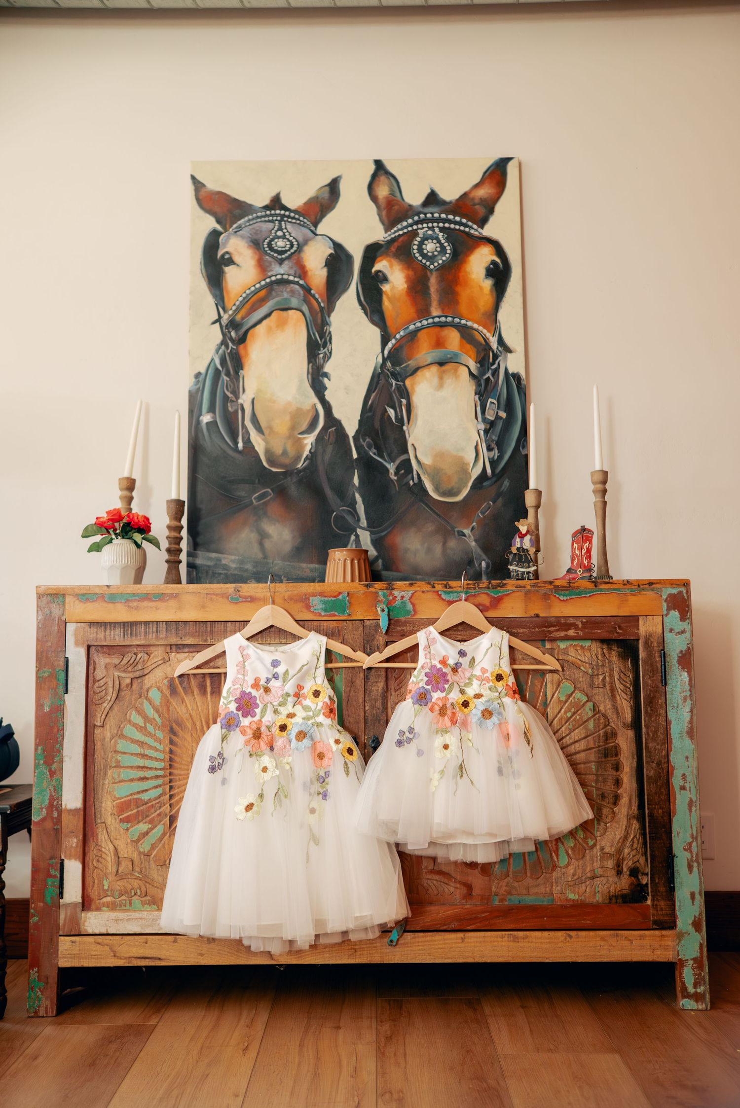 Floral flower girl dresses displayed on hangers against a rustic wooden backdrop with horse artwork.