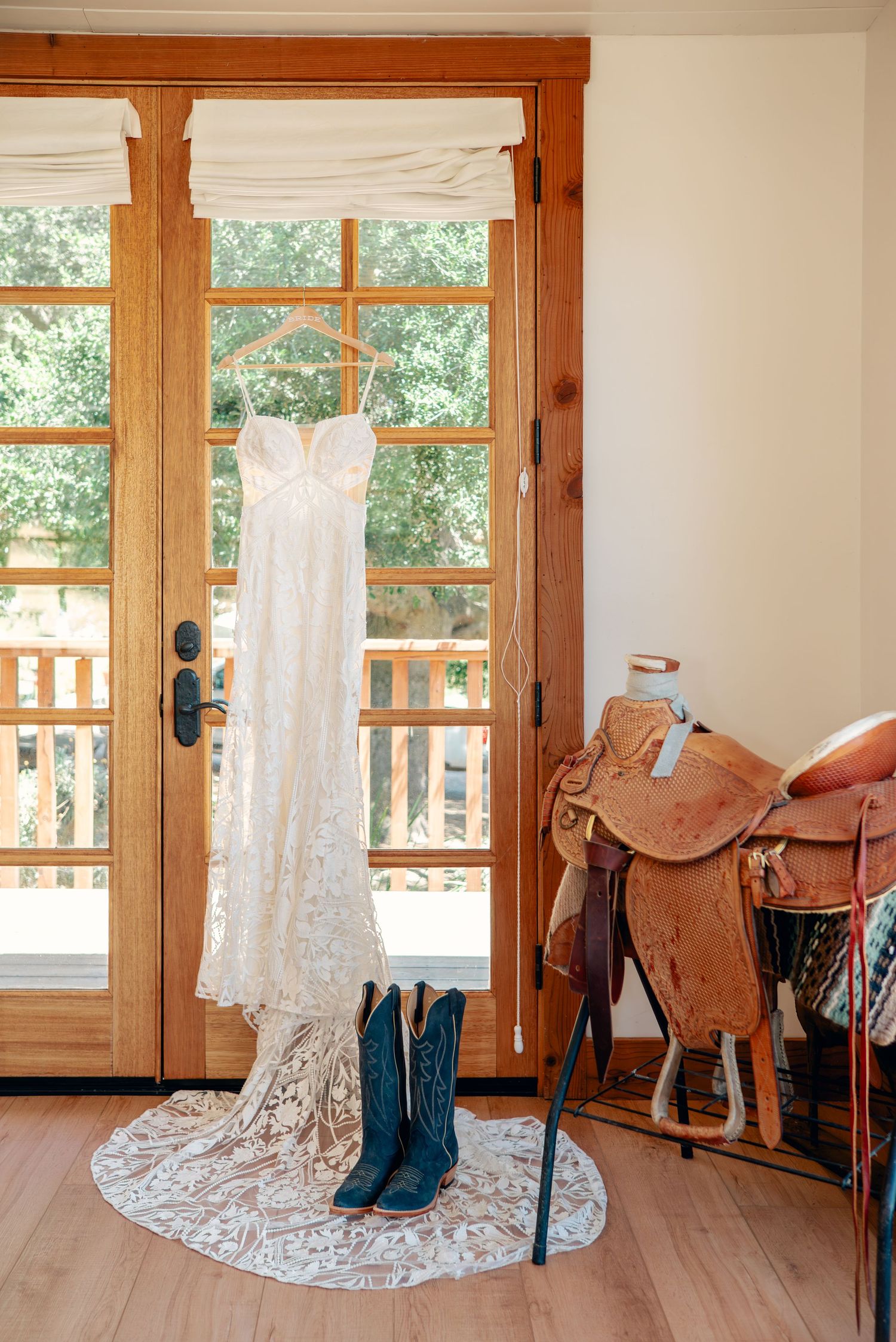 Elegant lace wedding dress displayed in a rustic room with wooden doors and leather saddles.