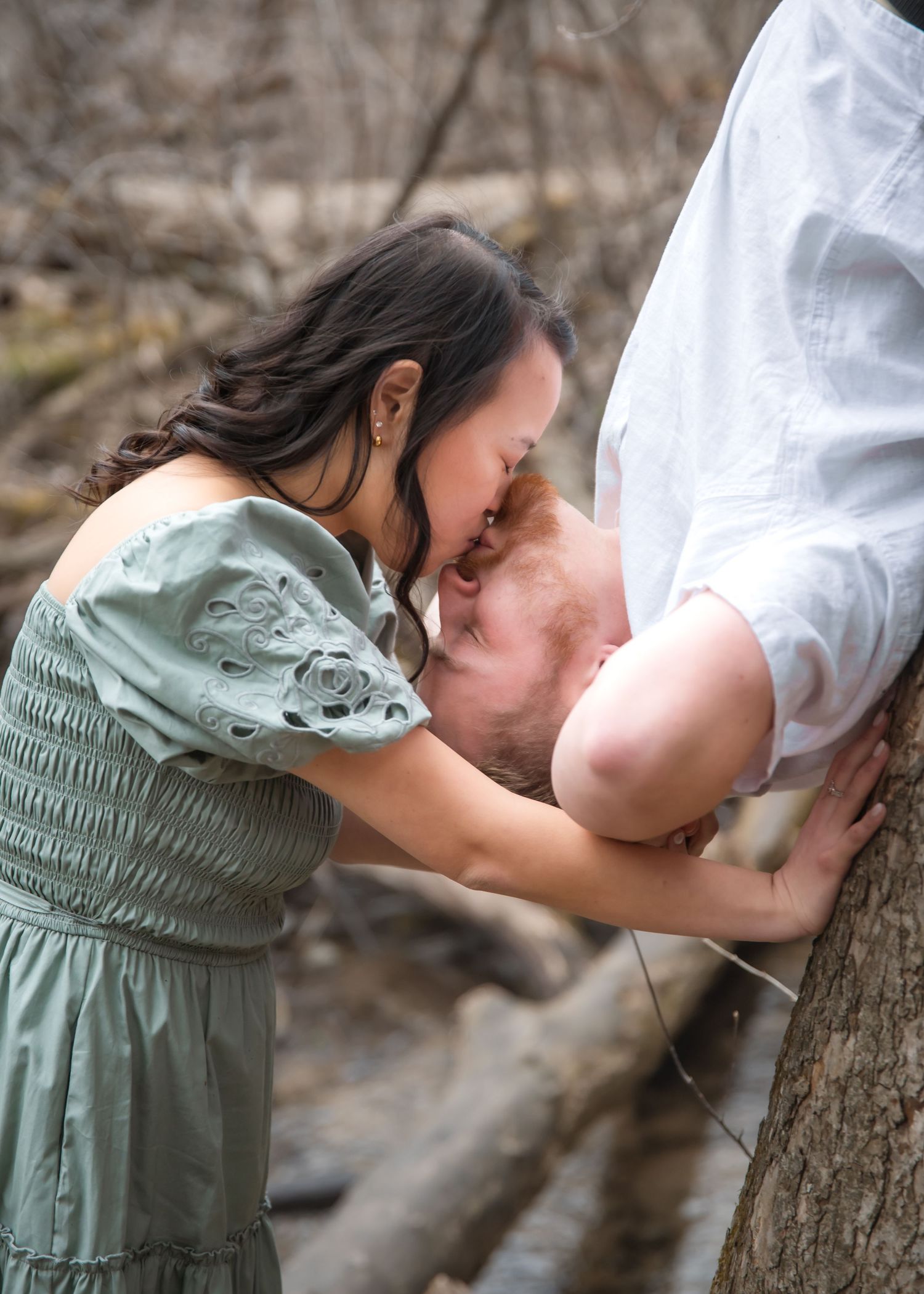 A couple shares intimate moments while sitting on fallen logs in a wooded area during an outdoor photoshoot.