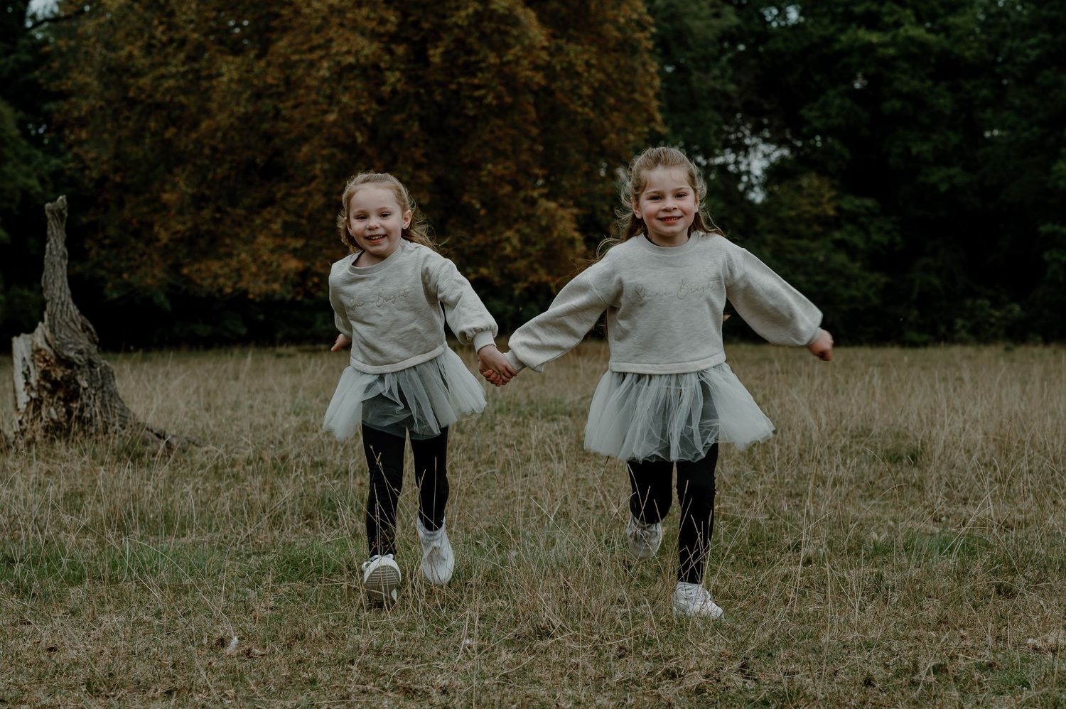 Two young children wearing matching gray sweaters and tutus run hand-in-hand through an autumn field.
