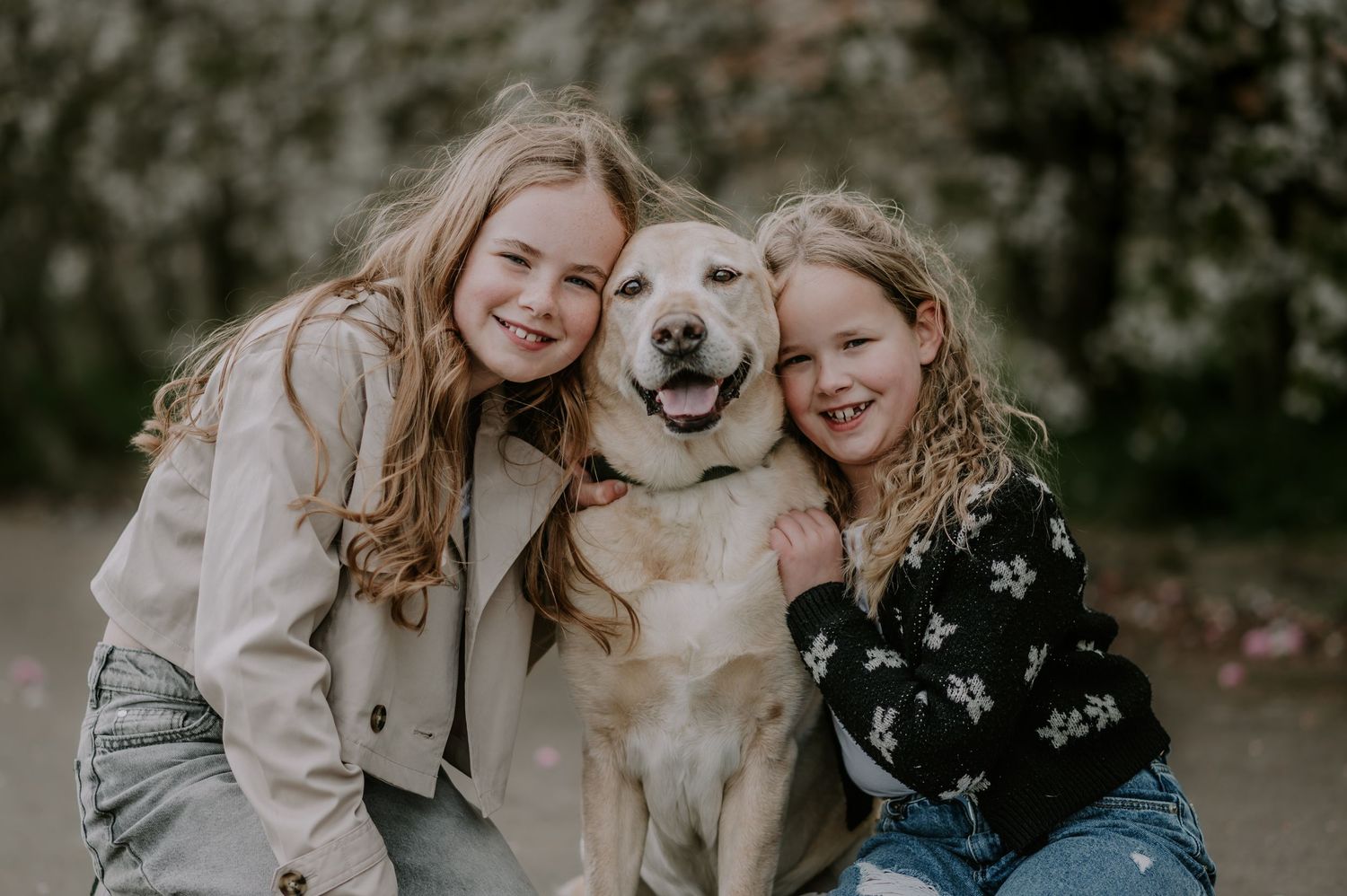 A happy yellow labrador dog smiles while being hugged by two people outdoors in a casual portrait setting.
