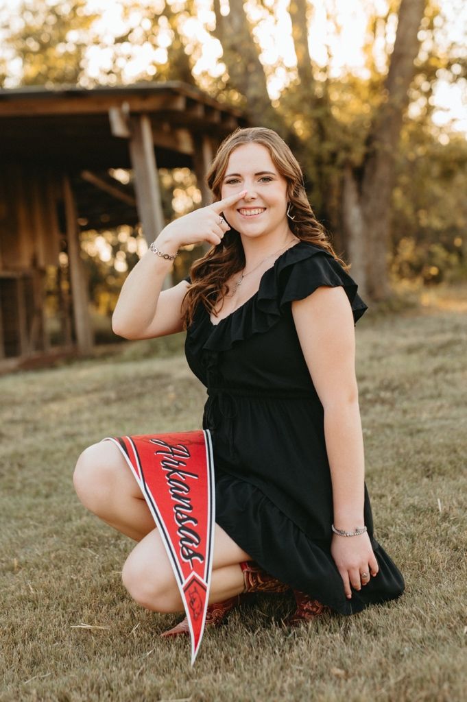 A cheerleader in a black top sits on grassy hill at sunset showing off red and white pom poms.