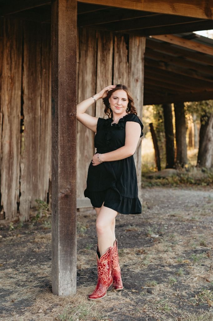 A person in a black dress and red cowboy boots poses playfully between wooden posts in a rustic outdoor setting.