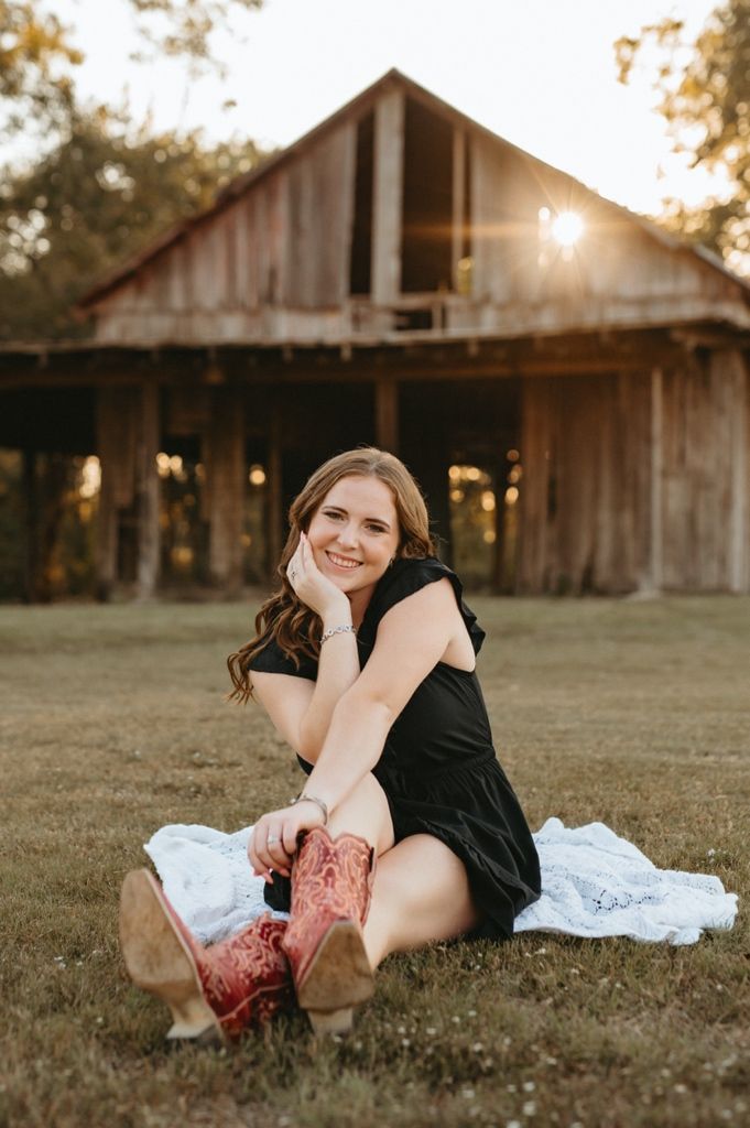 A person in black lounges on a white blanket in front of a rustic barn during golden hour sunset.