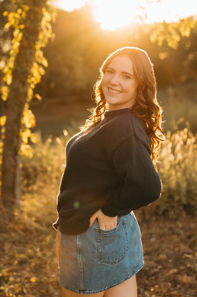 A woman in a black sweater and denim skirt poses outdoors during golden hour sunset with backlit glow.