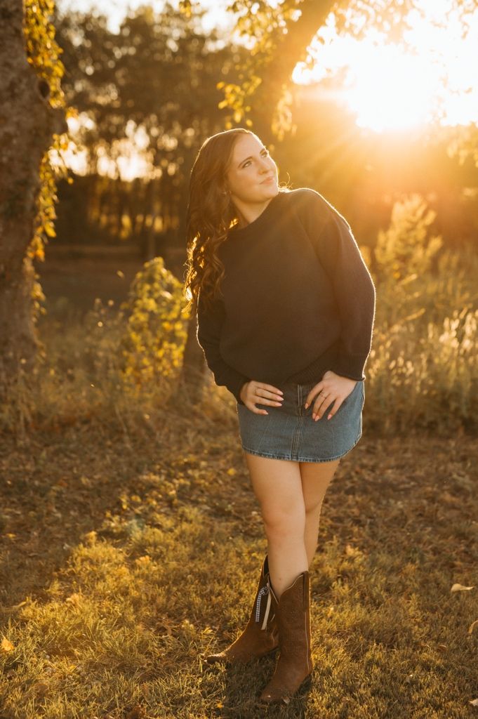 Person in black sweater and denim shorts poses in warm evening sunlight among trees.