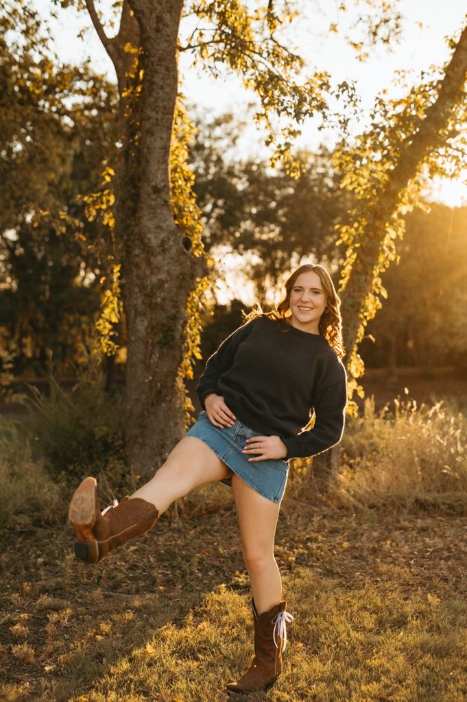 Person wearing denim shorts and black sweater poses playfully in a golden sunset forest setting.