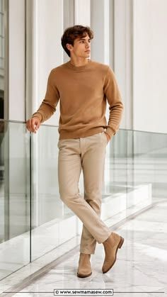 Man in a camel crewneck sweater with beige chinos and suede loafers, posing in a modern interior space.
