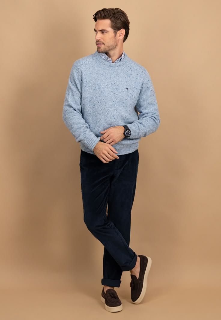 Man wearing a light blue speckled crew neck sweater with navy trousers and brown loafers against a beige background.