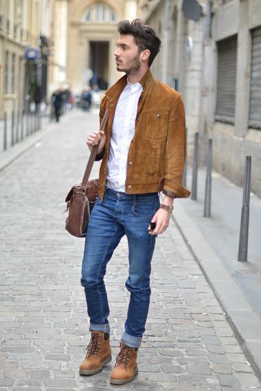 Stylish casual outfit featuring brown suede jacket, blue jeans, leather boots and messenger bag on cobblestone street.