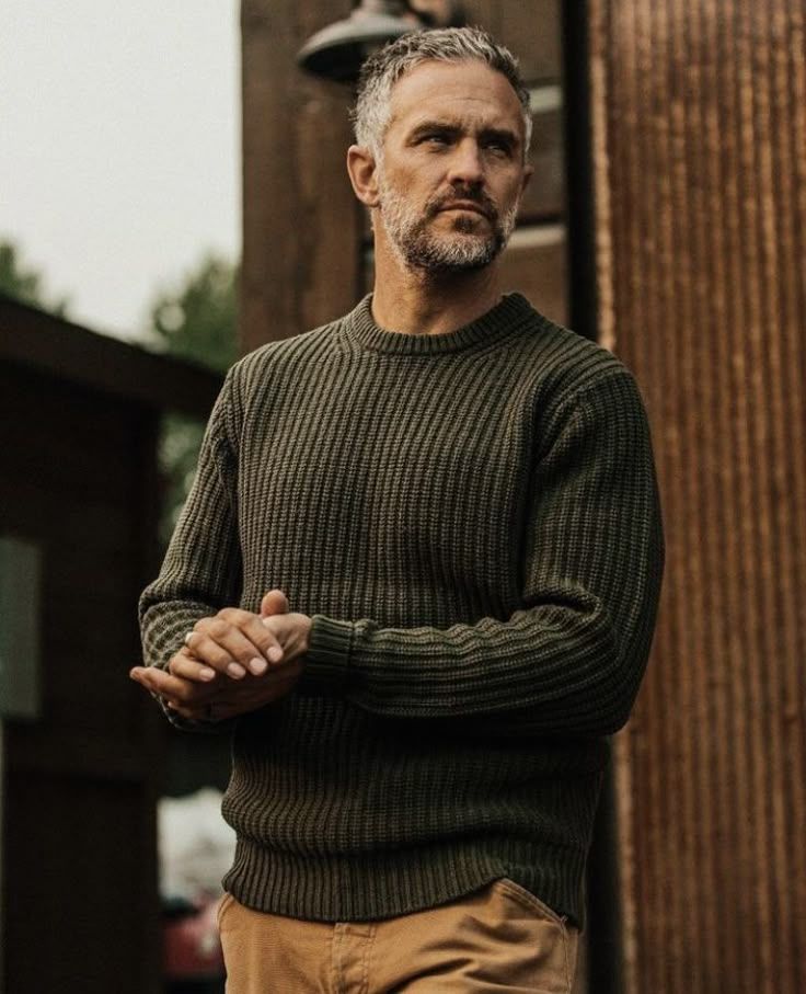 Man wearing olive green wool sweater stands confidently against wooden pillars in warm outdoor lighting.