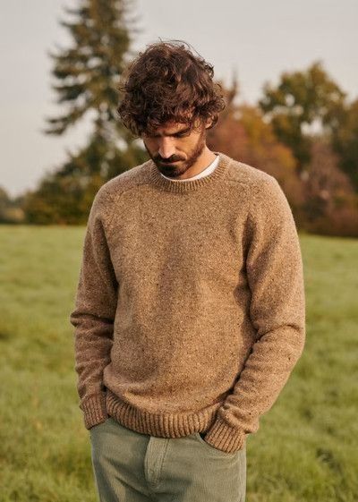 A person wearing a brown knit sweater poses outdoors in a green meadow during autumn.