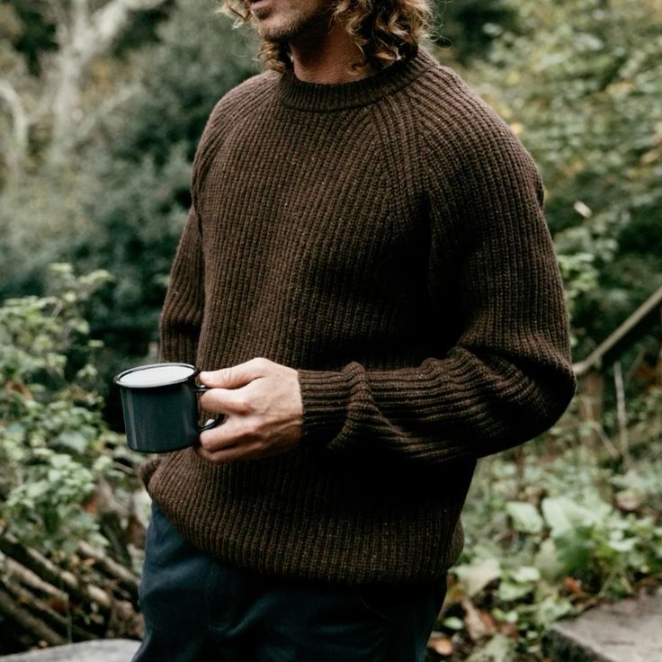 Person wearing brown knit sweater holding a dark mug while standing in a lush green forest setting.