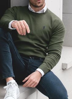 Man in olive green sweater and jeans sits casually by window with white sneakers.