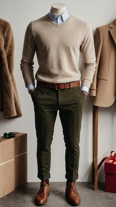 Stylish neutral men's outfit with beige sweater, blue shirt, brown leather shoes and dark pants displayed on white wall.