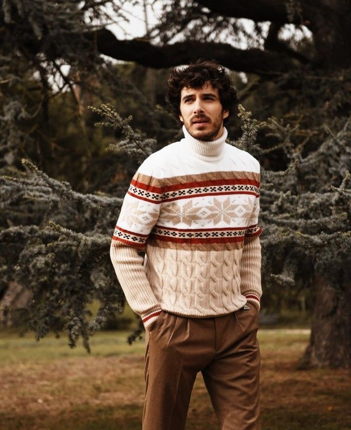 Man wearing a cream Fair Isle turtleneck cable-knit sweater with brown trousers, standing outdoors near pine trees.