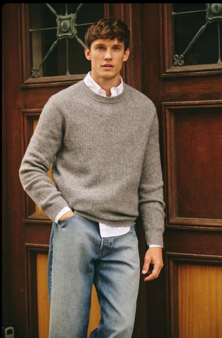 Man in gray knit sweater layered over white shirt with light blue jeans standing by wooden door.