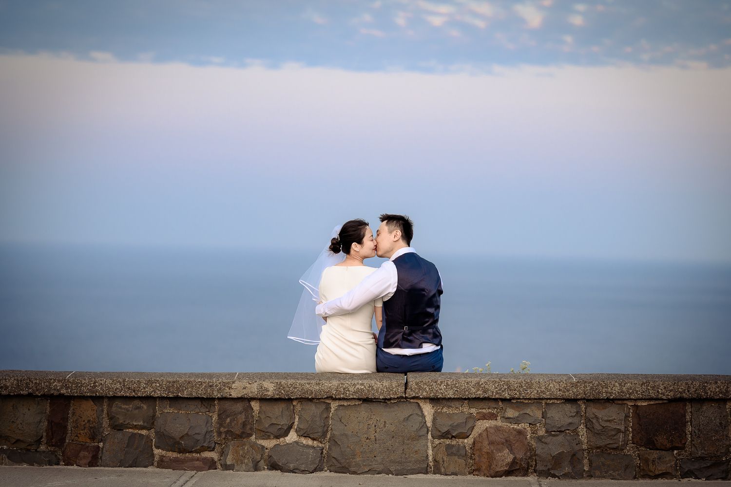 Newlywed couple sitting on a rock fence at Signal Hill and kissing with the Atlantic Ocean in the background.