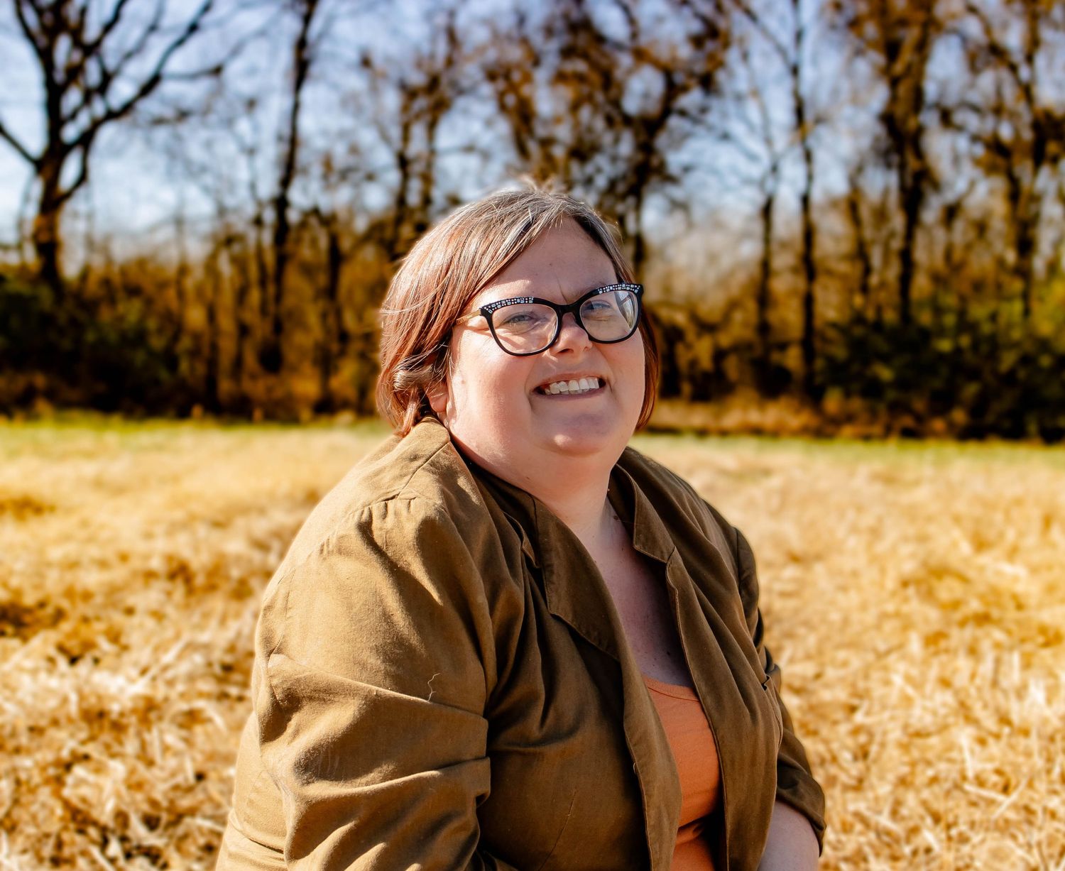A person wearing a brown jacket sits in a field of golden autumn leaves during sunset.