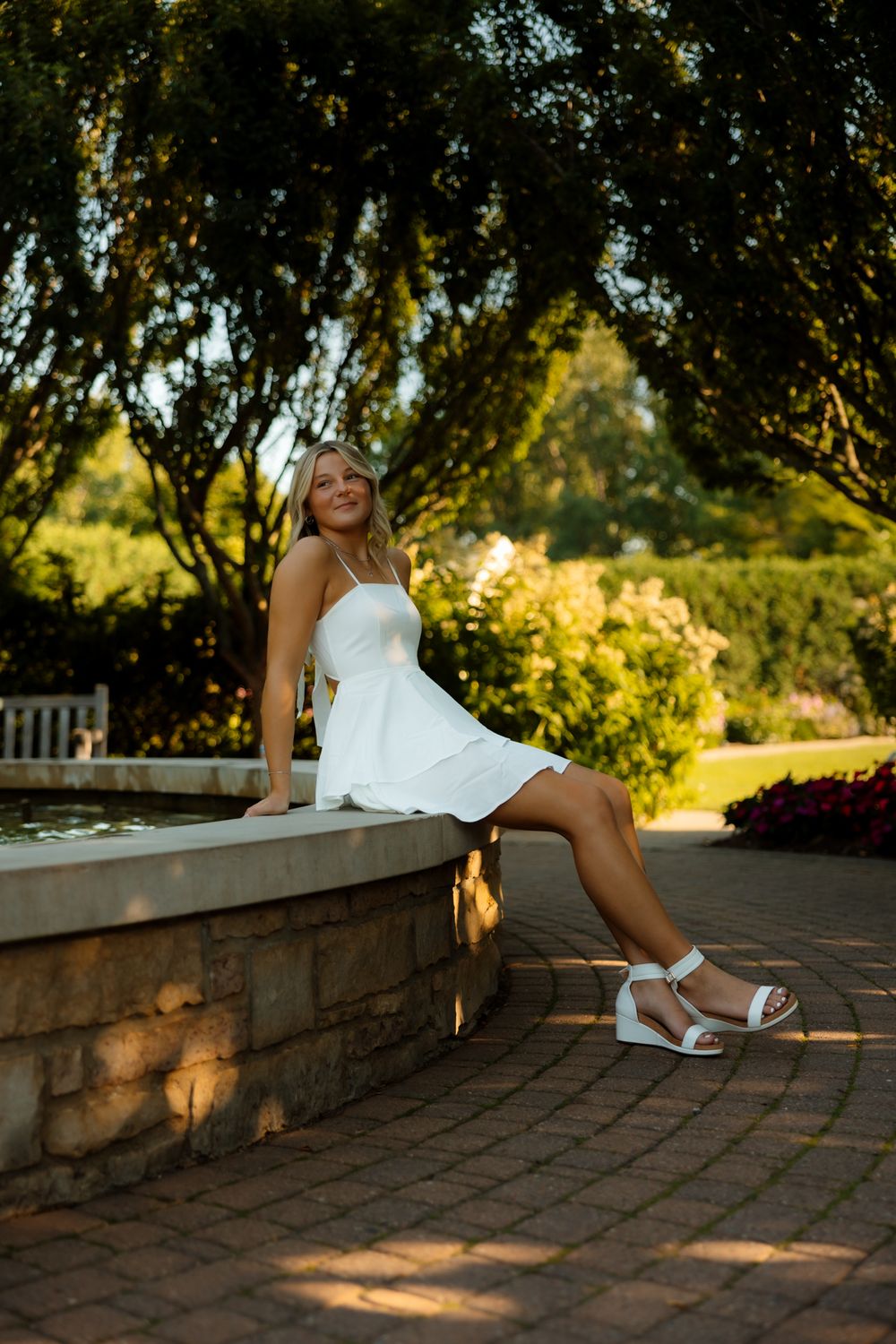 A person in a white sundress sits on a stone wall in a garden during golden hour lighting.