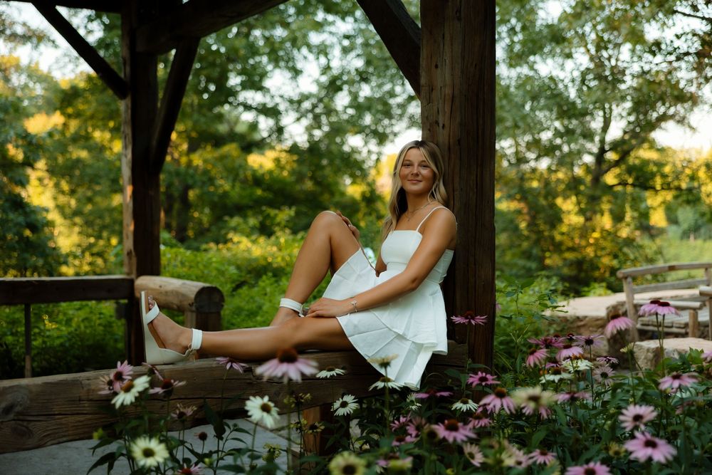 Person in white dress lounges on rustic wooden bench surrounded by blooming purple flowers at sunset.