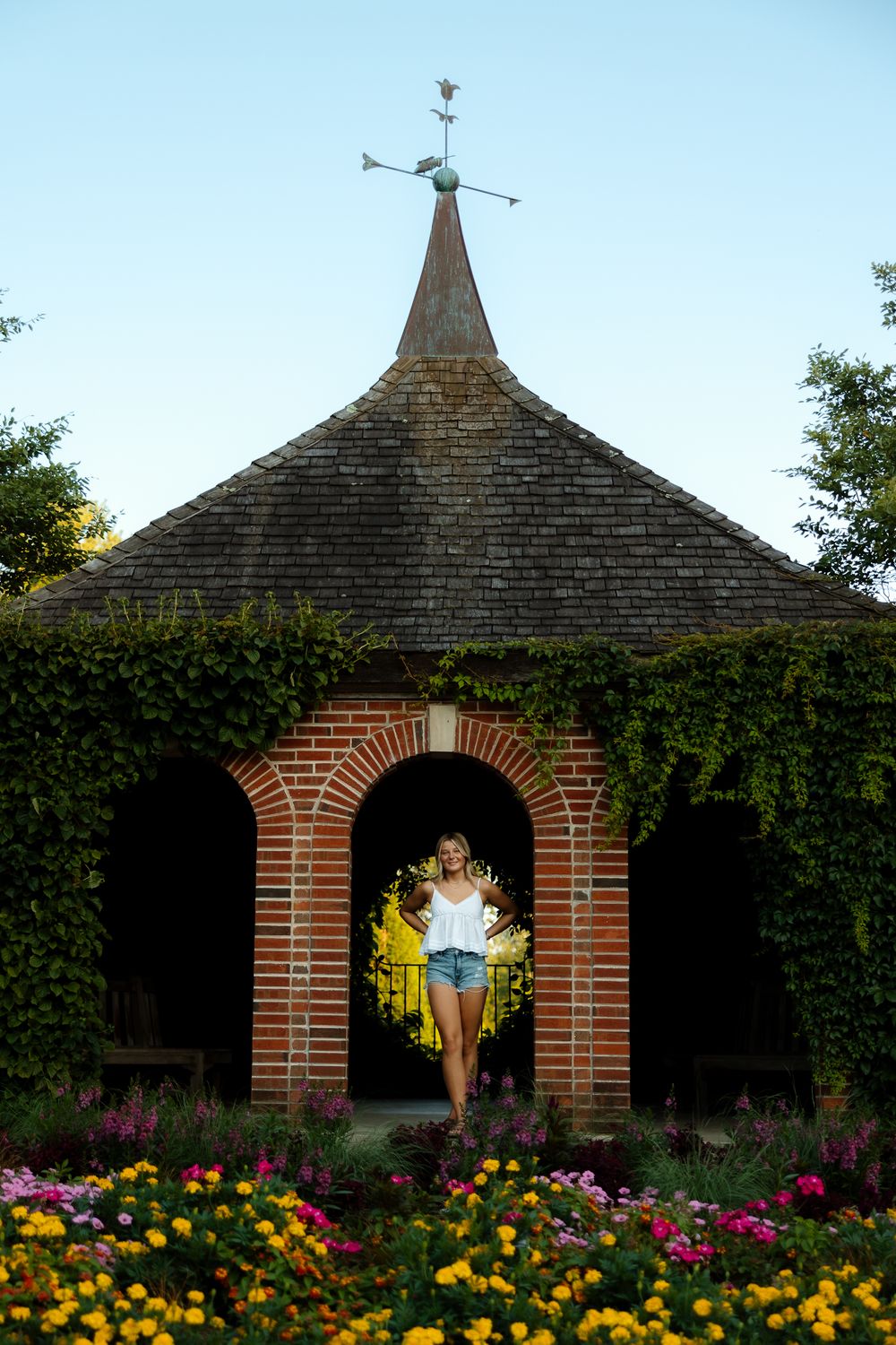 Brick archway with wooden shingle roof surrounded by colorful flowers in an English garden setting.
