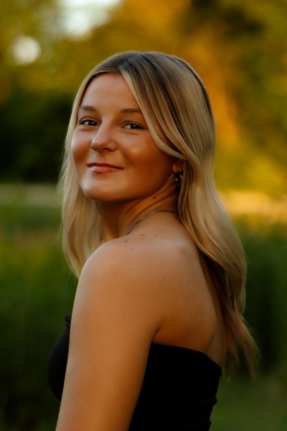 Portrait of someone with blonde hair wearing a black strapless top against a warm sunset background with bokeh effects.