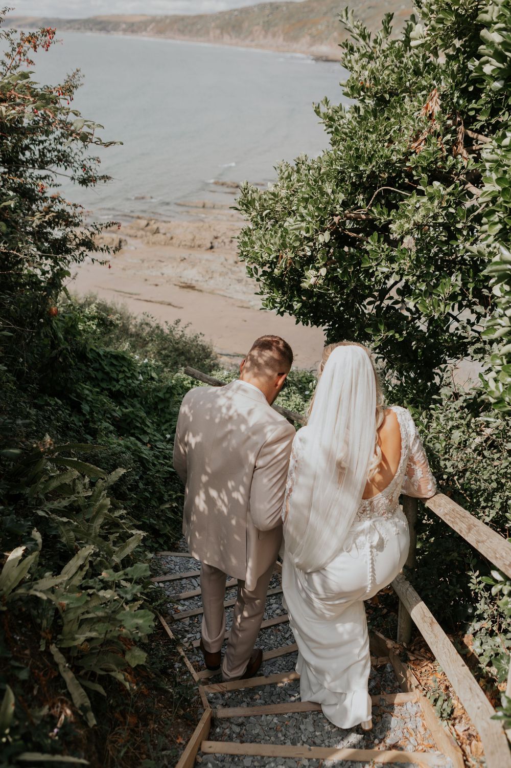 Bride and groom descend wooden coastal steps toward a scenic beach on their wedding day.
