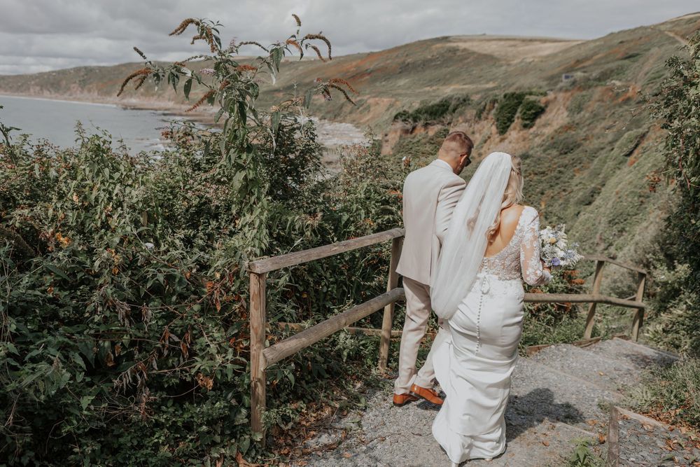 Newlyweds hold hands on a clifftop path with stunning coastal scenery behind them.