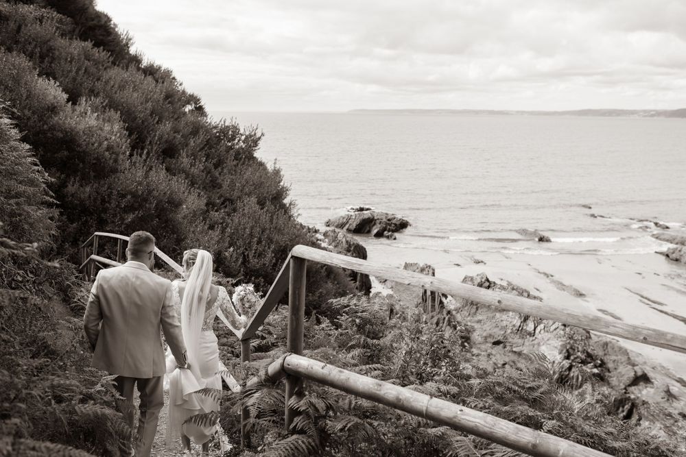 Bride and groom walk hand in hand down coastal steps toward the sea in black and white.