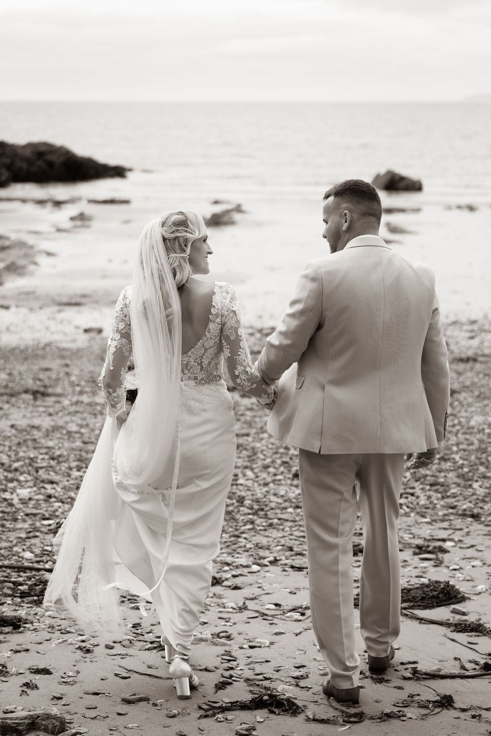 Bride and groom walk hand in hand along a rocky beach in black and white wedding photo.