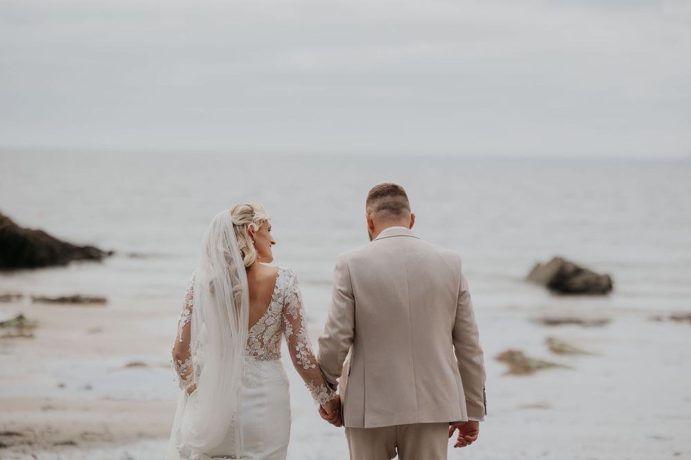 Bride in lace gown and groom in tan suit walk hand in hand along an overcast beach.