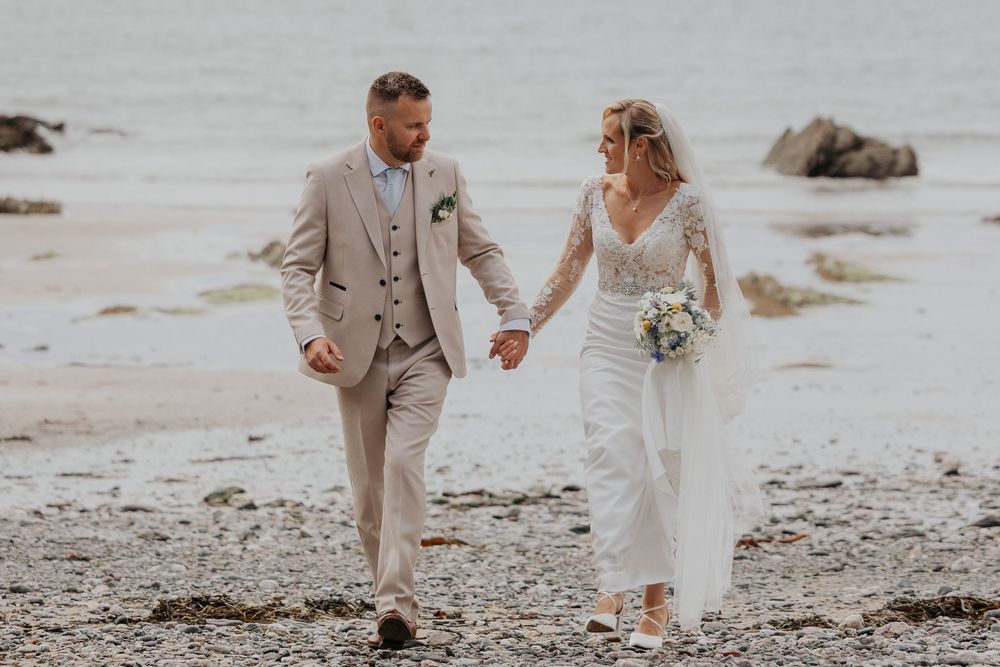 Newlyweds smile and hold hands while strolling along a rocky beach on their wedding day.