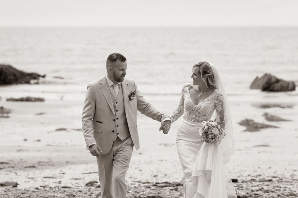Bride and groom hold hands and gaze at each other on a rocky beach in black and white.