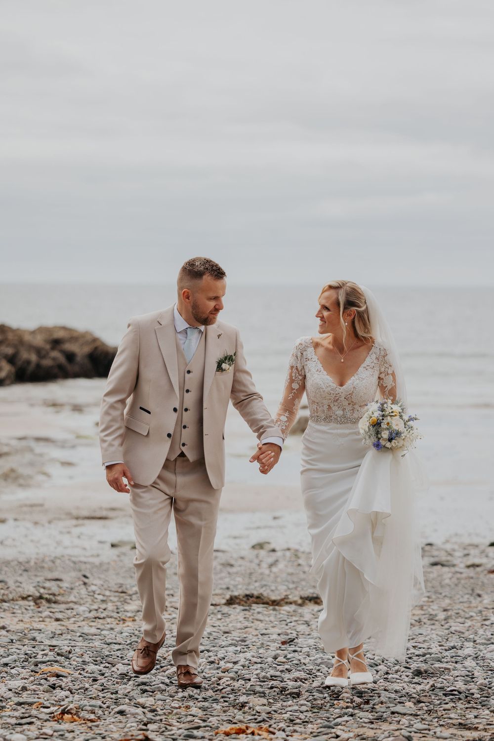 Bride and groom walk hand in hand along a pebble beach smiling at each other.