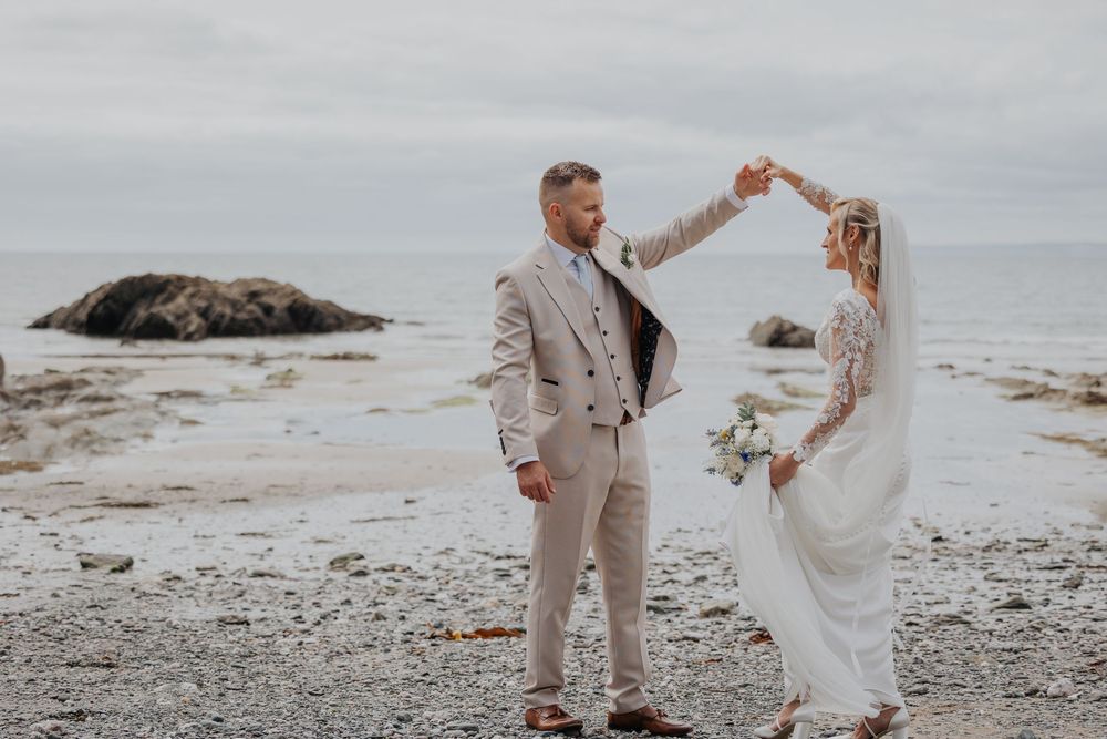 Groom in beige suit twirling bride in lace wedding dress on rocky beach with overcast sky.