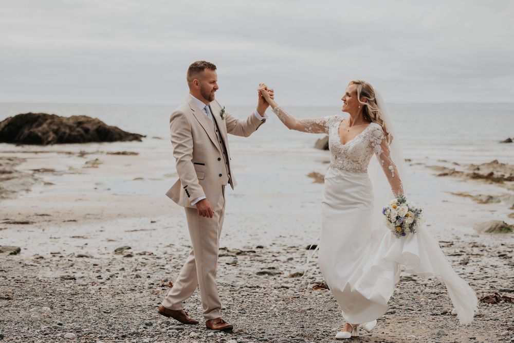 Bride and groom dancing hand in hand on a pebble beach, bride holding a blue and white bouquet.