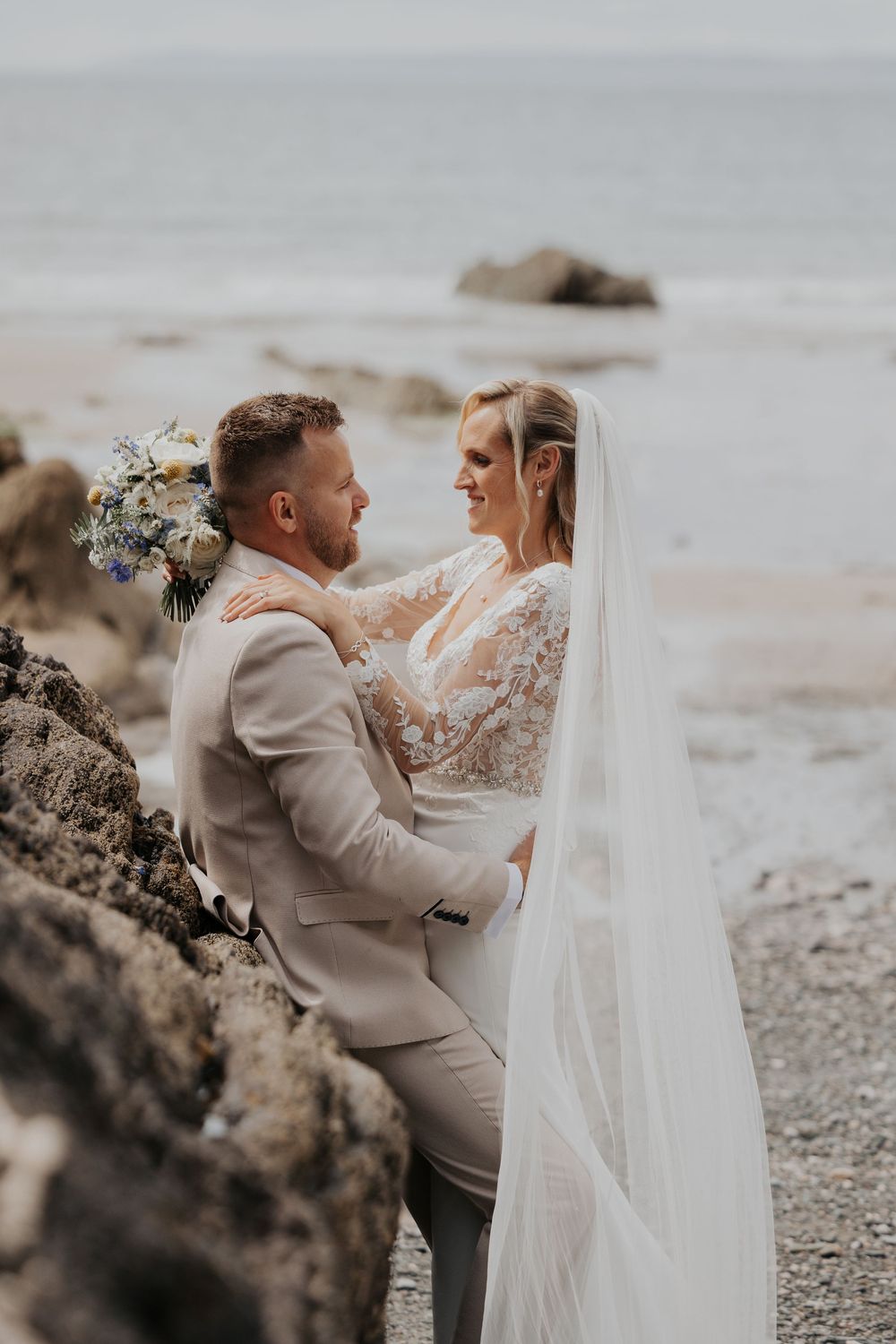 Bride and groom sharing a romantic moment by coastal rocks, bride in lace gown with flowing veil.