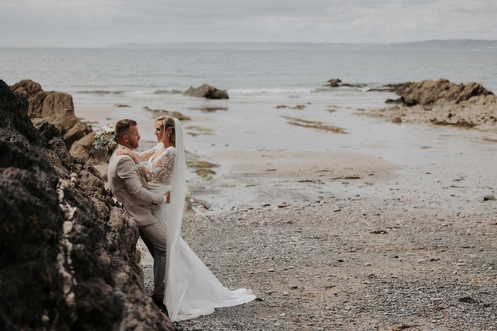 Bride and groom standing close together by seaside rocks on a cloudy beach wedding day.