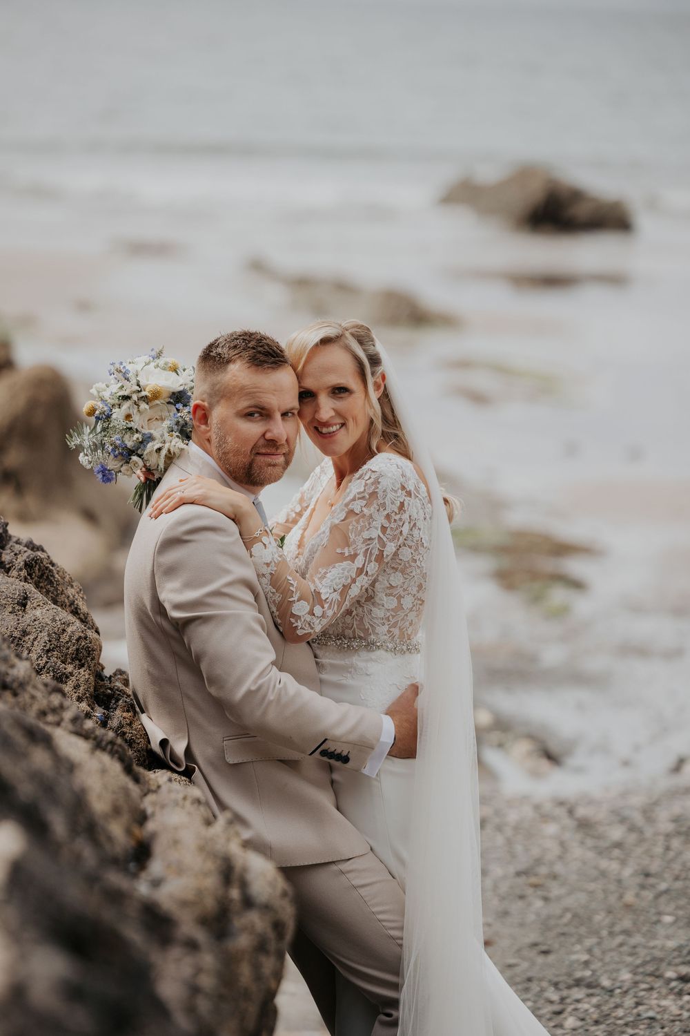 Bride embracing groom from behind on rocky beach, holding a blue and white floral bouquet.
