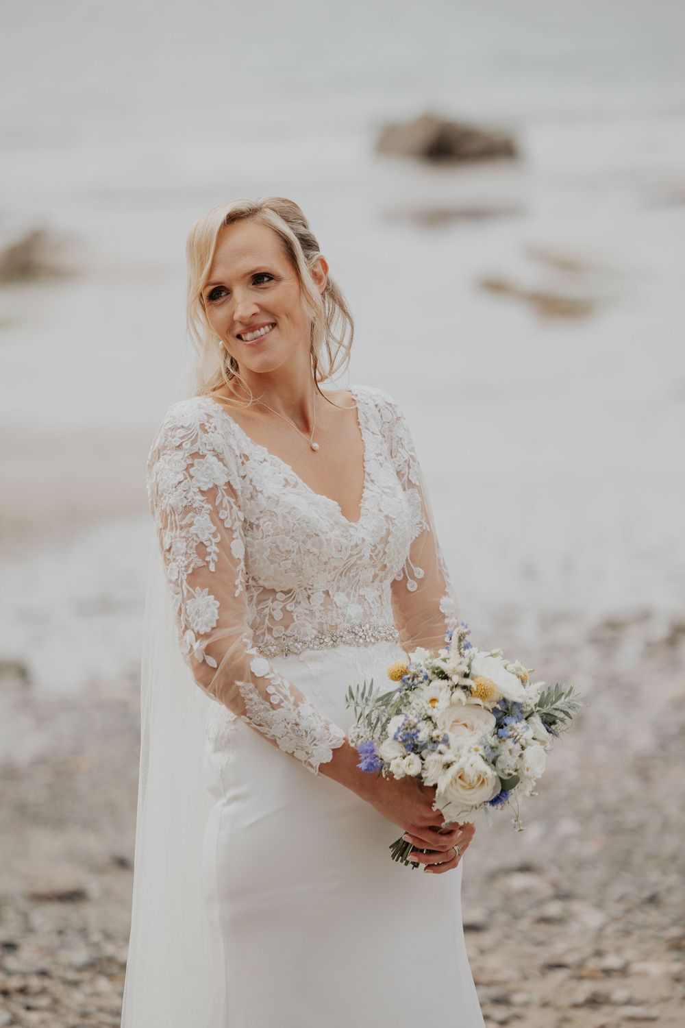 Smiling bride in lace long-sleeve wedding gown holding a blue and white bouquet on a beach.