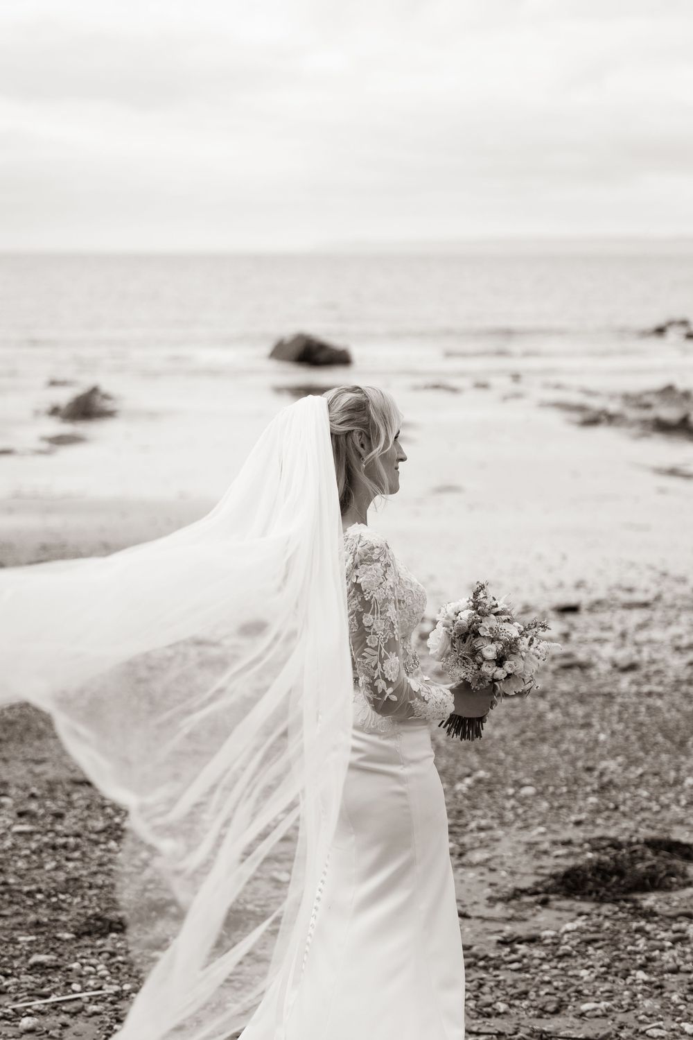 Black and white photo of bride with flowing veil blowing in wind on a rocky beach.