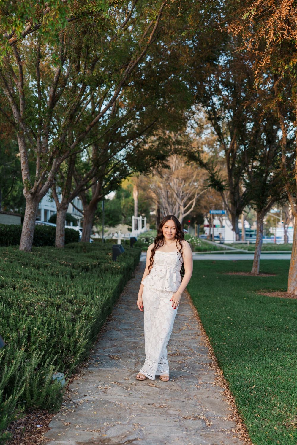 Walking along a tree-lined autumn path wearing a white lace dress with fallen leaves scattered around.