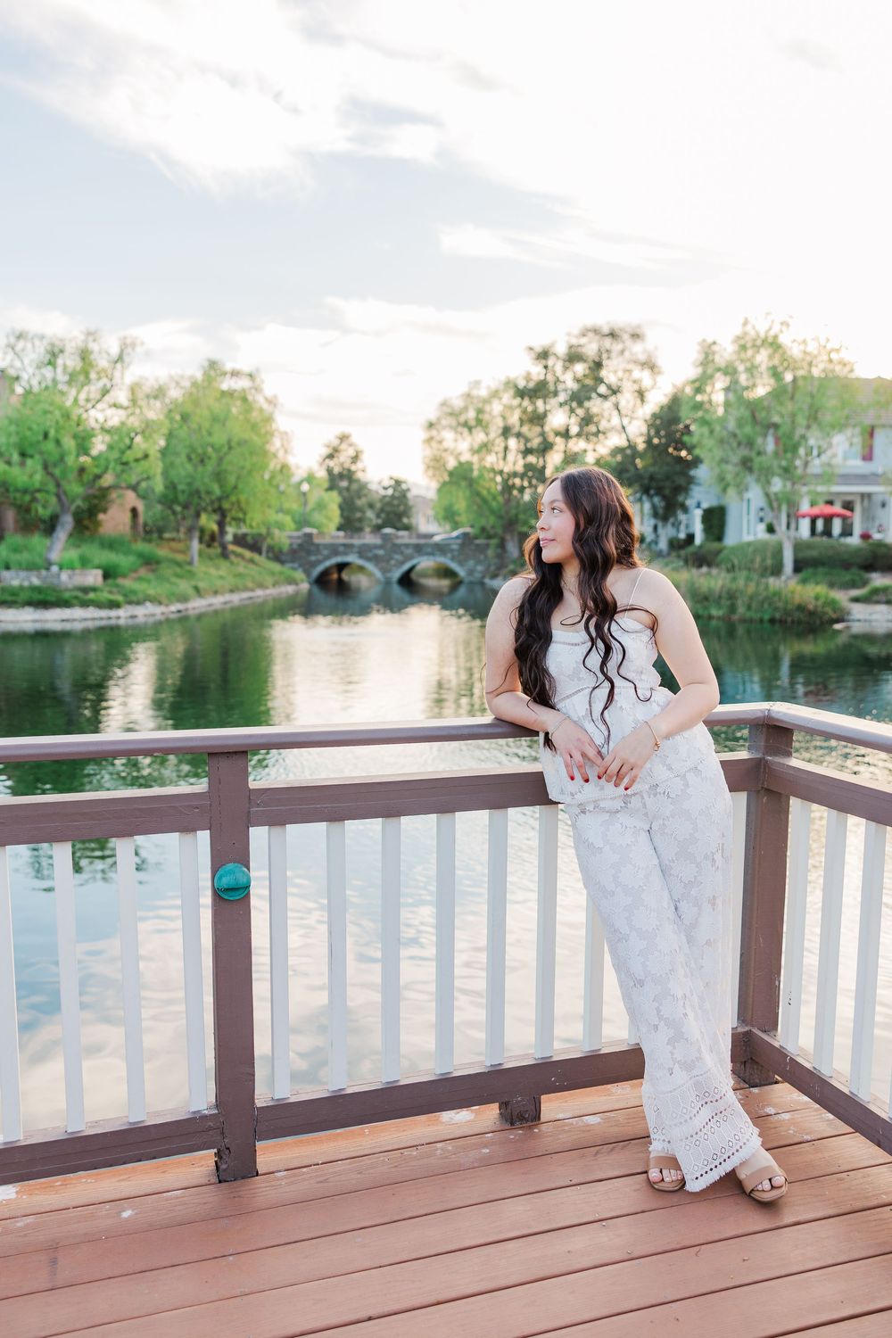 A woman in an elegant white dress stands on a wooden deck overlooking a tranquil pond with a stone bridge in the background.