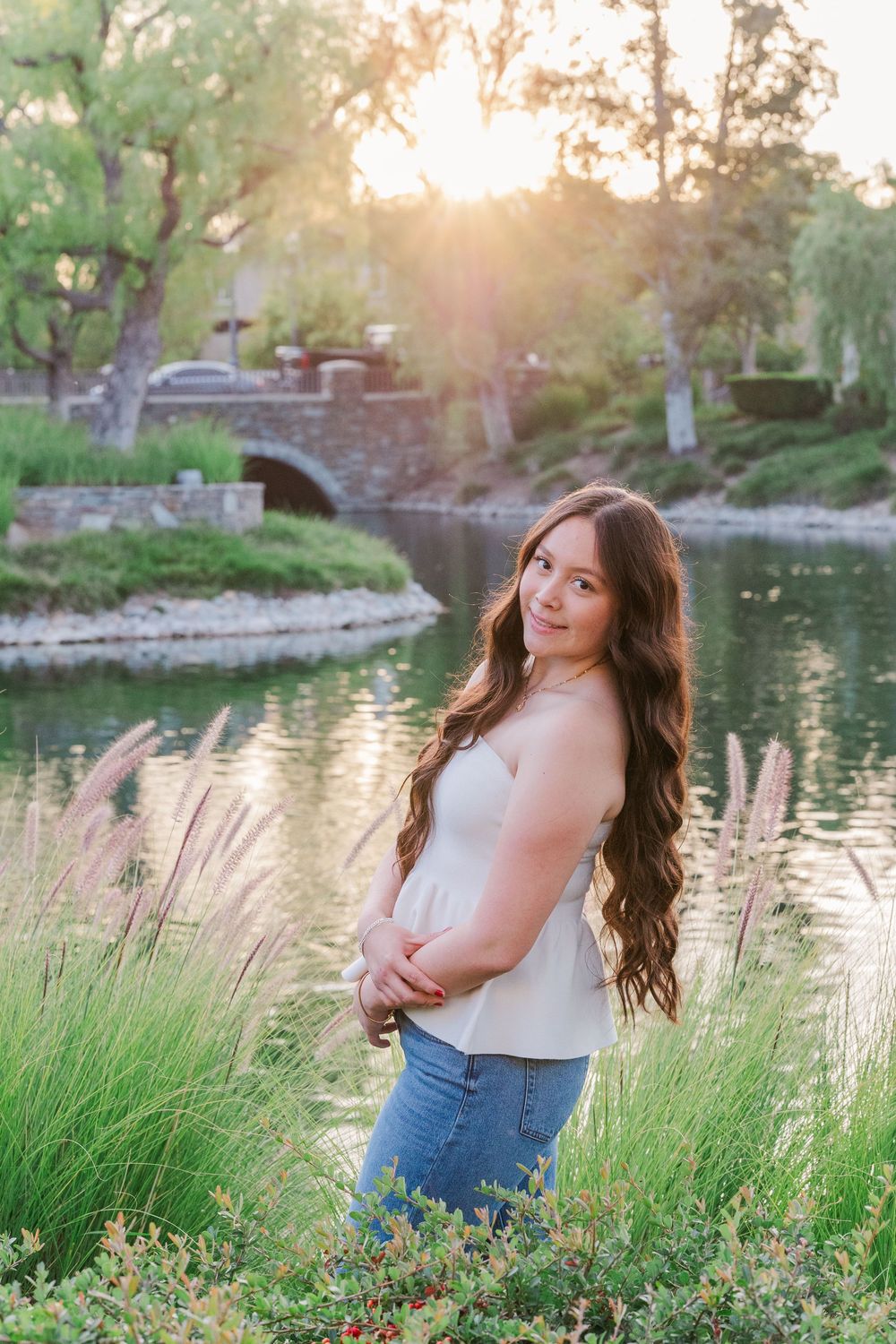 A woman in casual attire poses near ornamental grasses by a pond during golden hour with a stone bridge in the background.