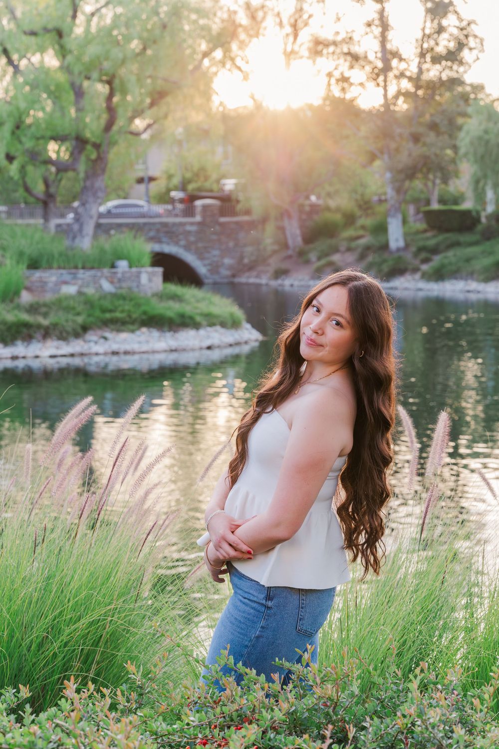 A woman in a white tank top and jeans poses playfully in tall grass by a pond during golden hour sunset.